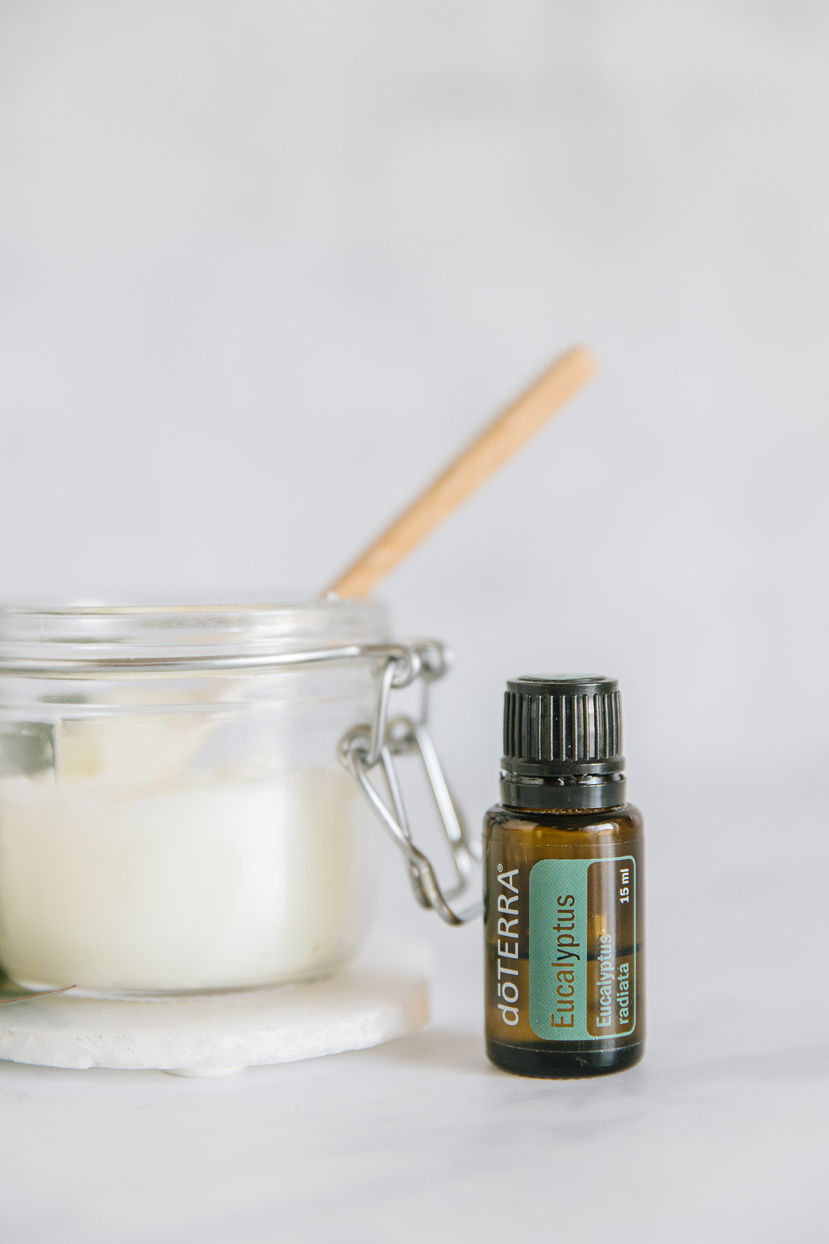 A glass jar with a wooden spatula inside of it on top of a white coaster with a Eucalyptus Doterra essential oil next to it.