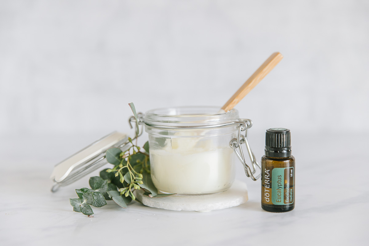 A glass jar with homemade vicks rub with a small spatula with a wooden handle inside of it with a green plant around it on top of a white coaster next to a Doterra Eucalyptus essential oil.