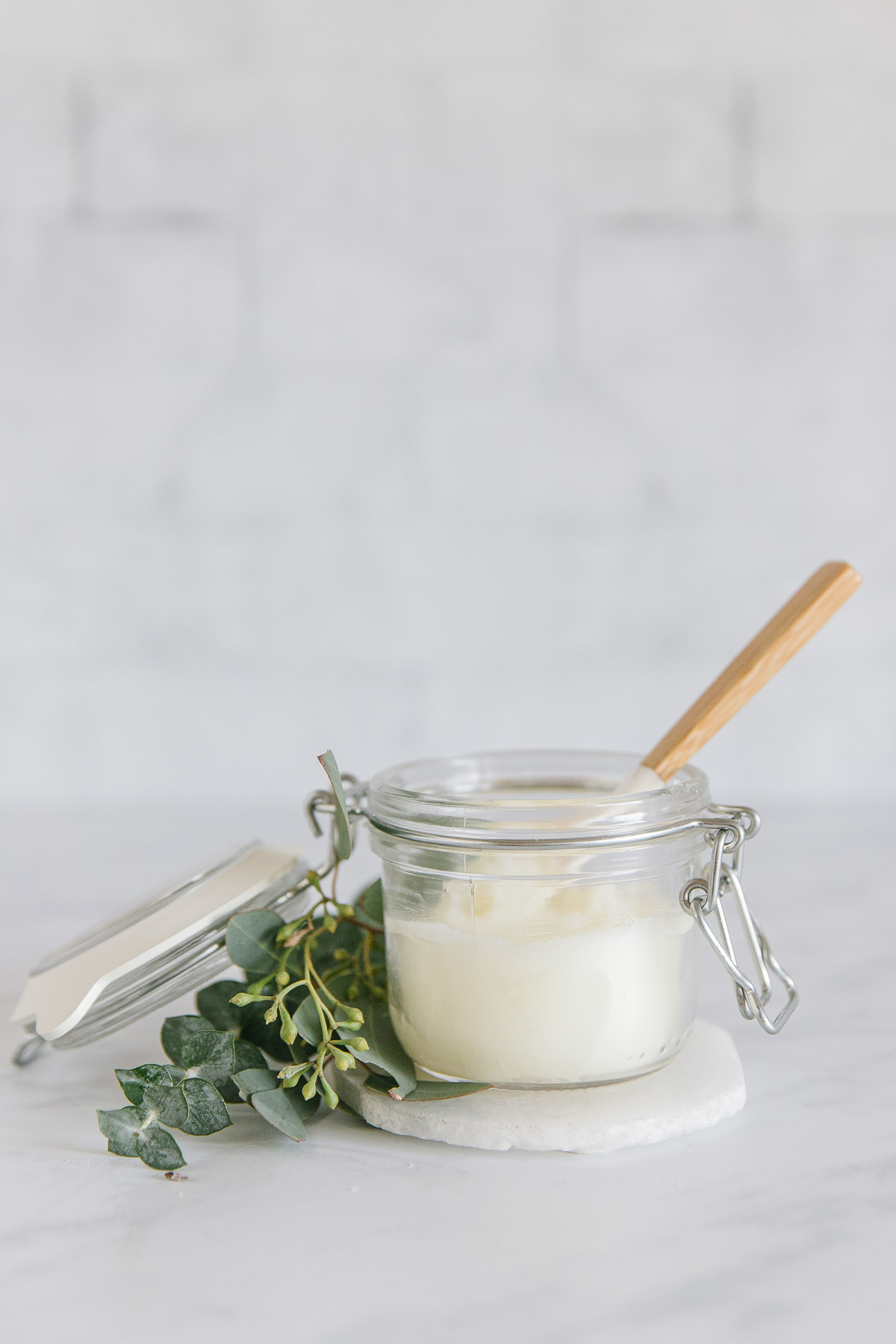 A glass jar with a small spatula with a wooden handle inside of it with a green plant around it on top of a white coaster.