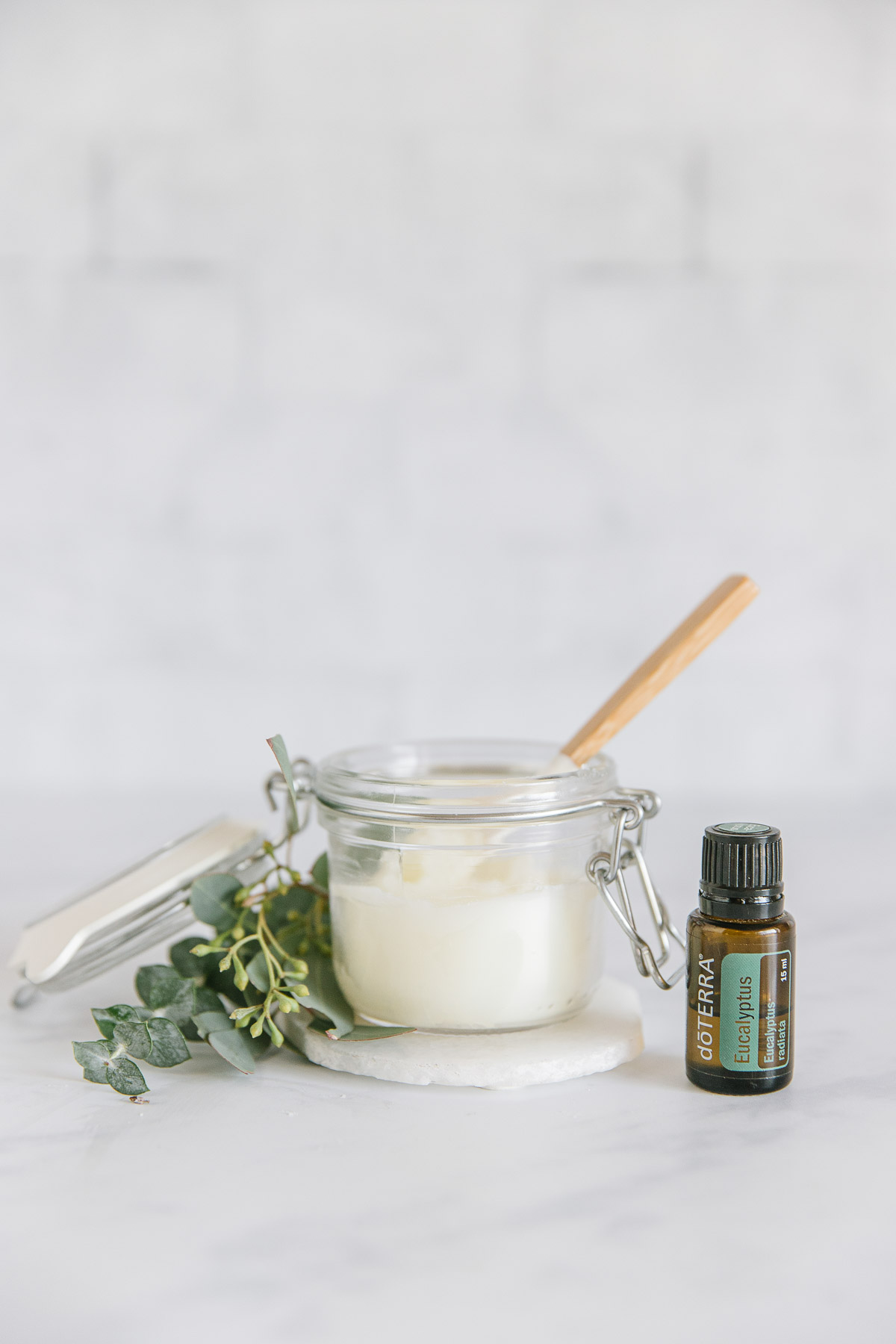 A glass jar with homemade Vicks vapor rub with a small spatula with a wooden handle inside of it with a green plant around it on top of a white coaster with a Eucalyptus Doterra essential oil next to it.