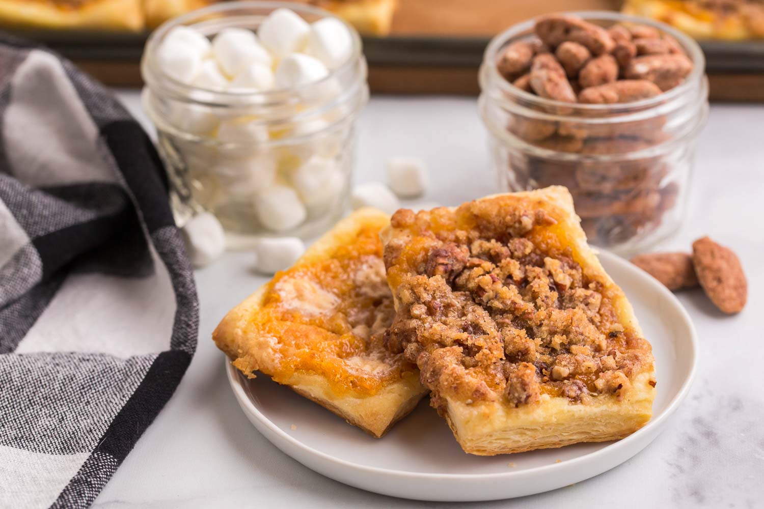 Two pieces of decadent pastry on a white plate with two glass jars filled with pecans and marshmallows.