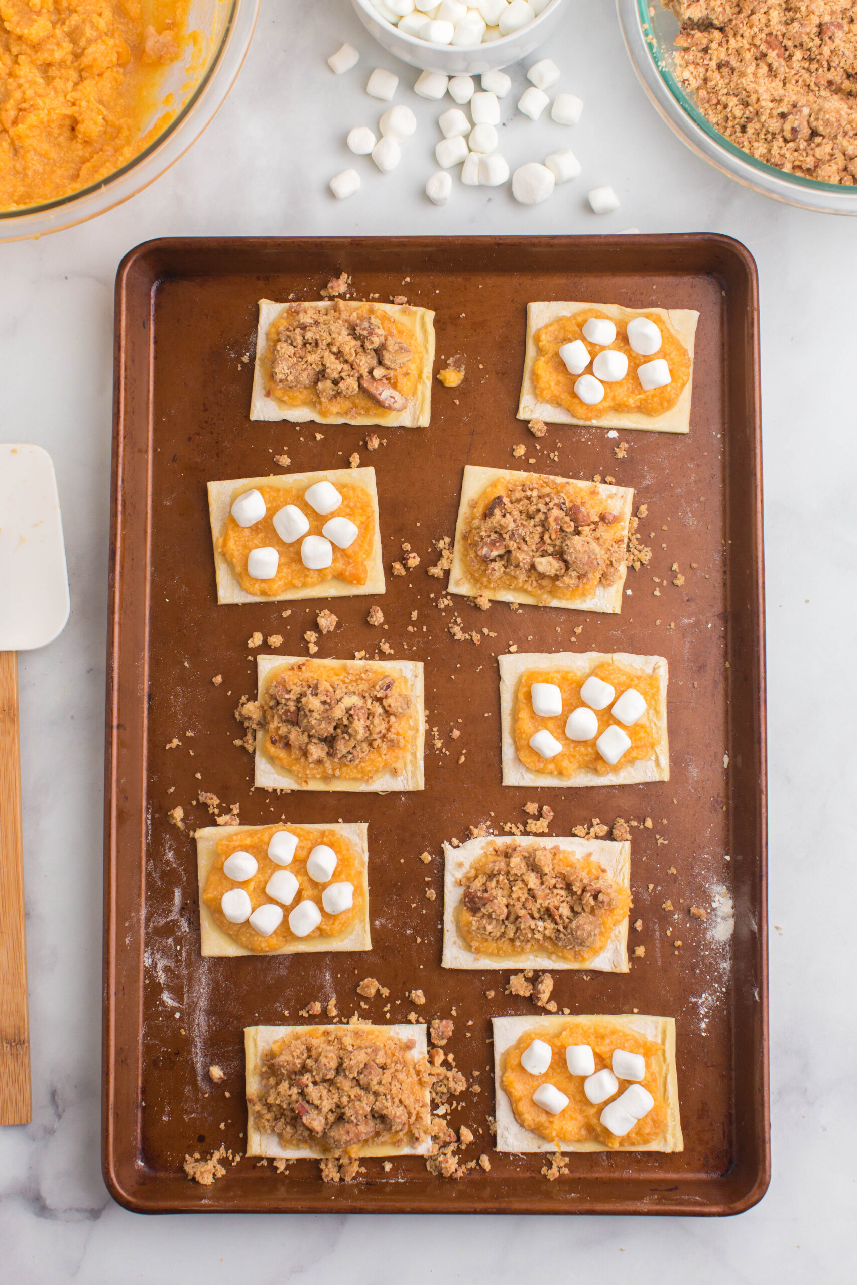 A copper pan with eight unbaked pastry with toppings. There is a white spatula and extra ingredients of brown sugar, marshmallows and sweet potatoes at the top.
