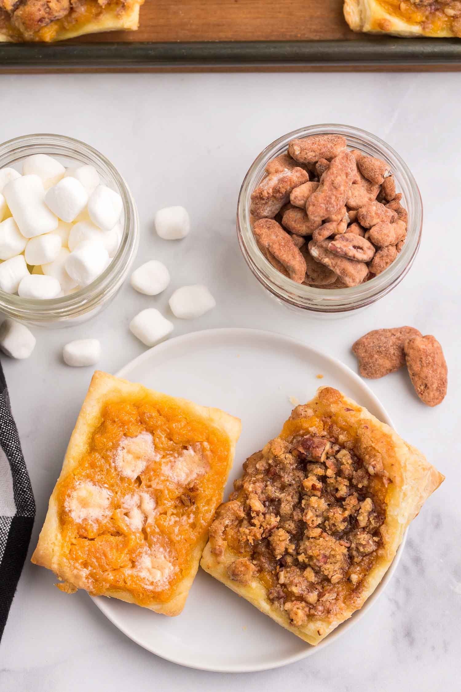 Two pieces of sweet potato pastry on a white plate with two cups of marshmallows and pecans and a plate filled with pastry behind it.