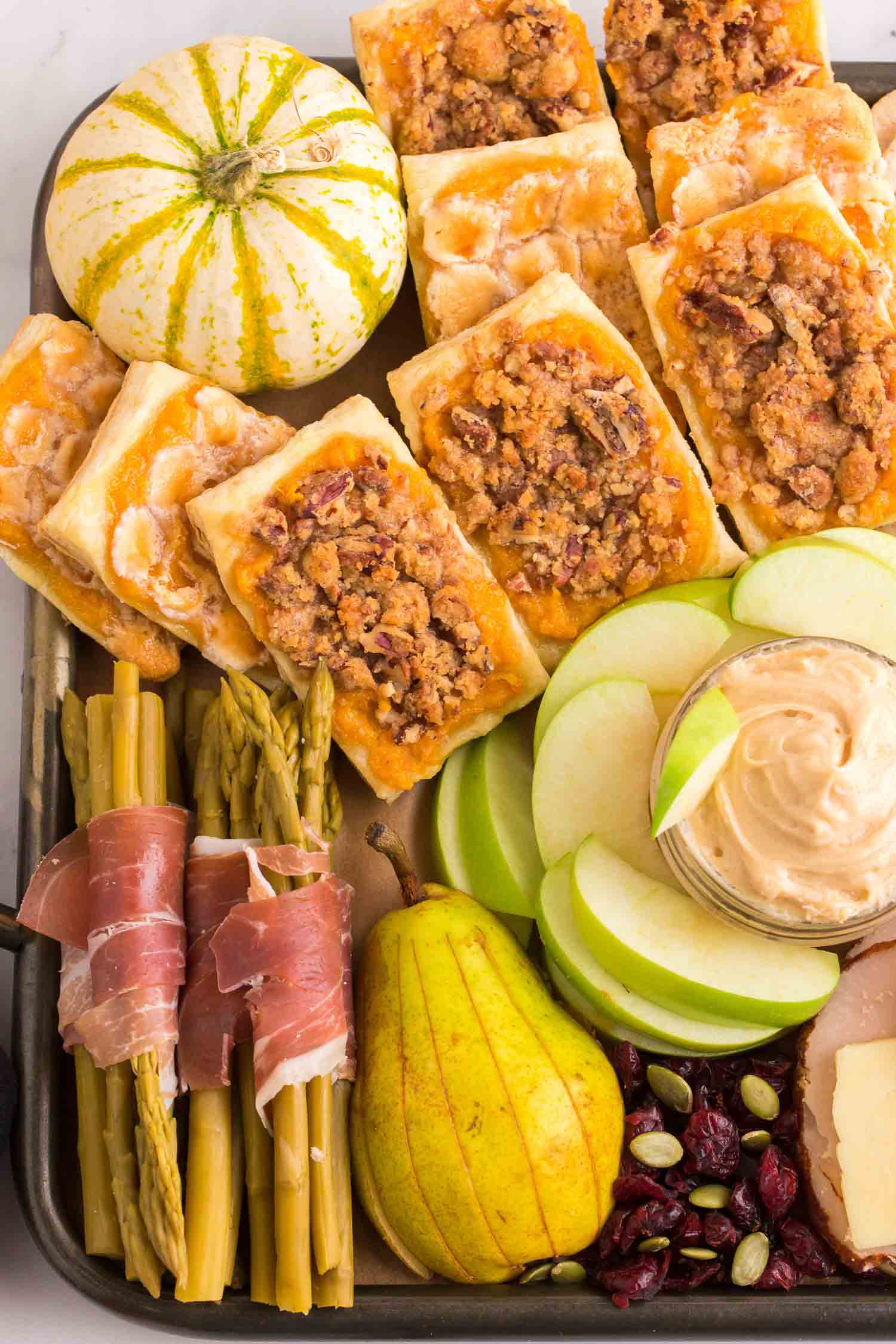 A platter with sweet potato pastries, a white pumpkin. meat wrapped asparagus, cut pear, cut apples and apple dip, and nuts.