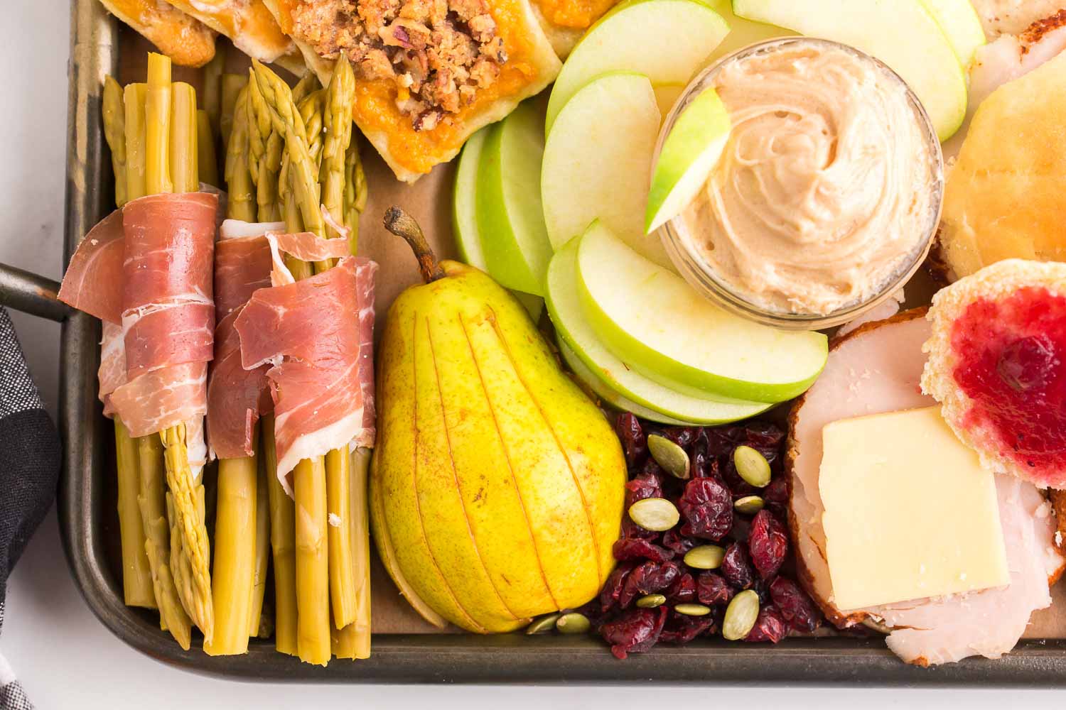Sweet potato pastries, meat wrapped asparagus, cut pear, cut apples and apple dip, nuts, and a roll with turkey and jelly on it.