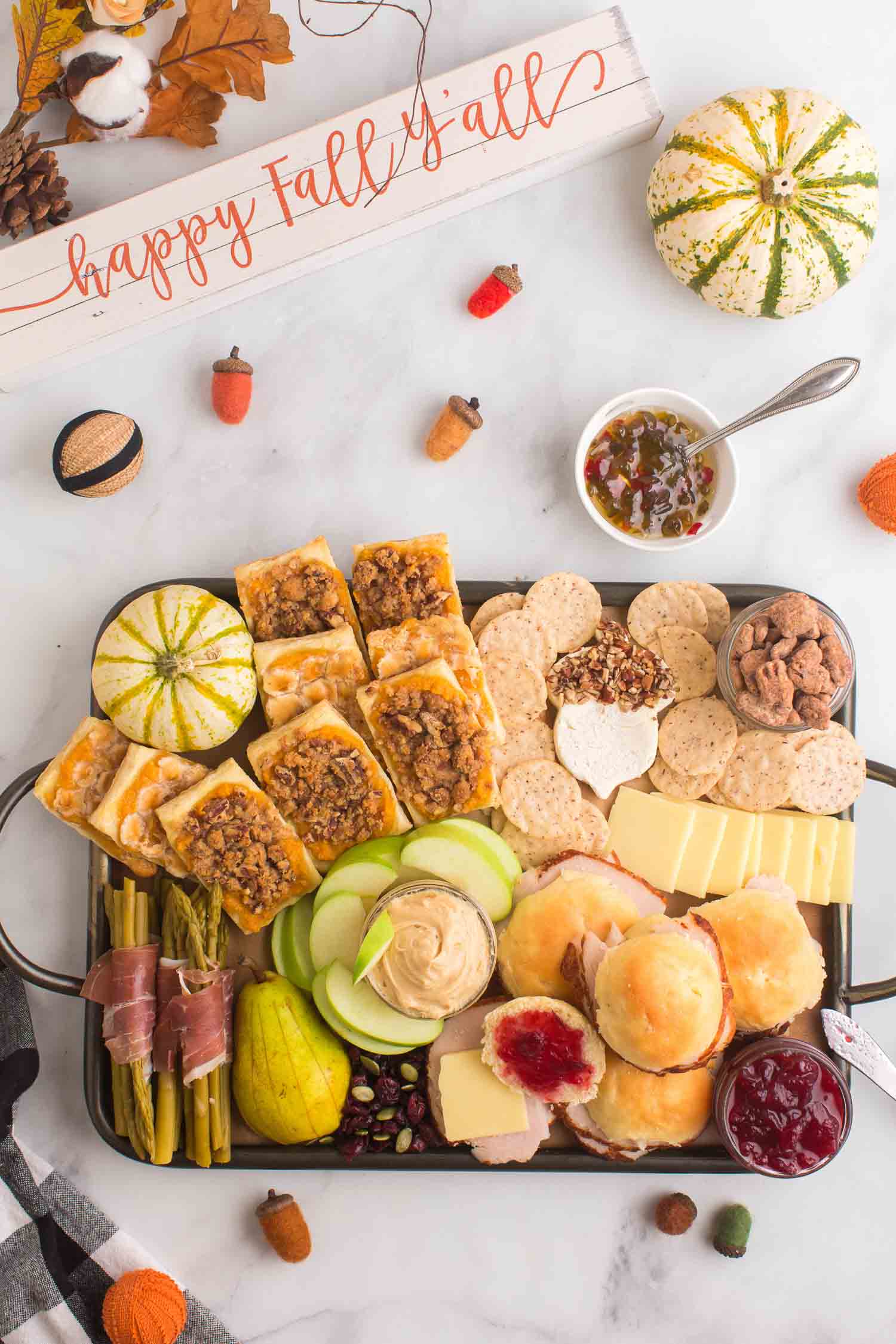 A full Thanksgiving charcuterie board with pumpkins, fruit, and other décor around the board and a bowl of pepper jelly with a silver spoon in it.