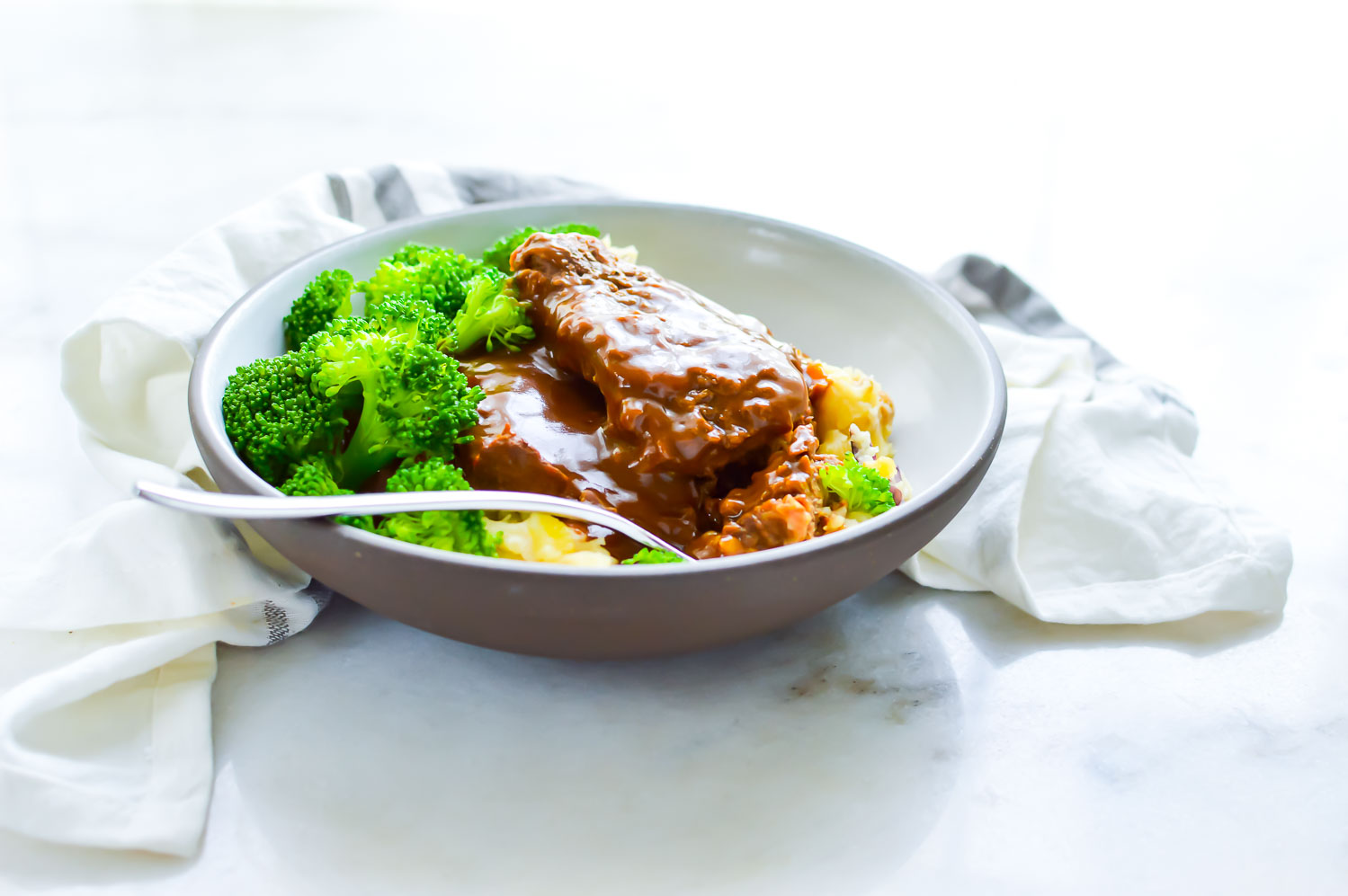 A white bowl with gray rim with meat, gravy, potato, and broccoli with a fork in it and a towel around it.