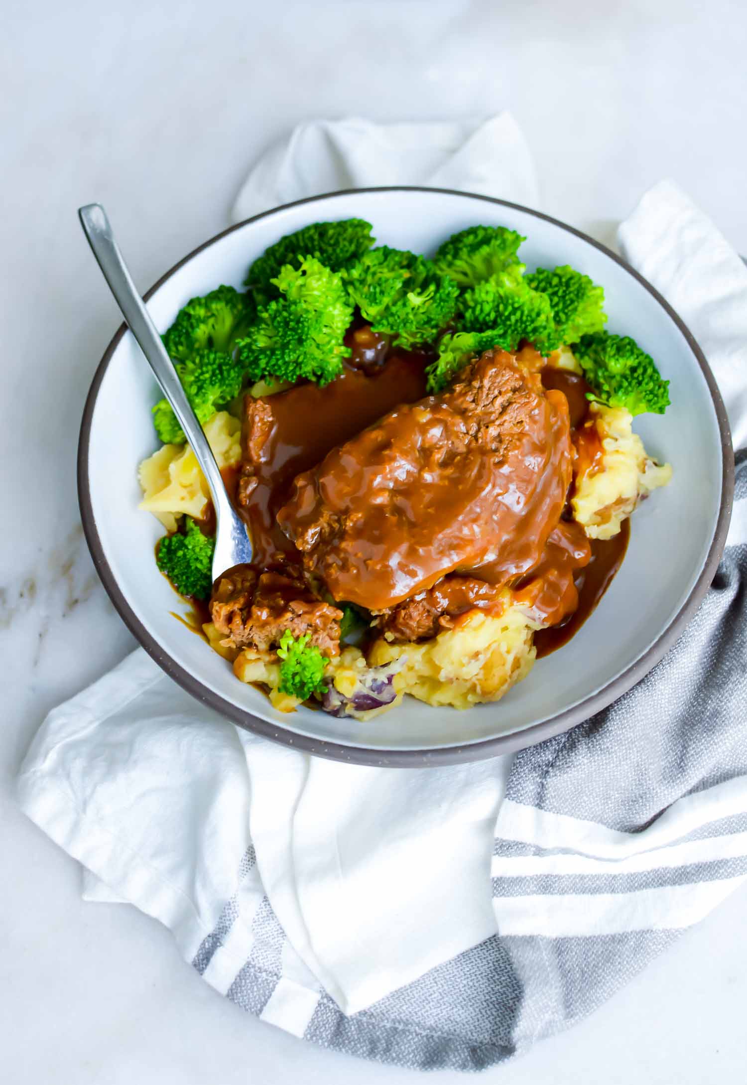 A white bowl with gray rim with cubed steaks, potato, gravy, and broccoli with a fork in it and a towel surrounding it.