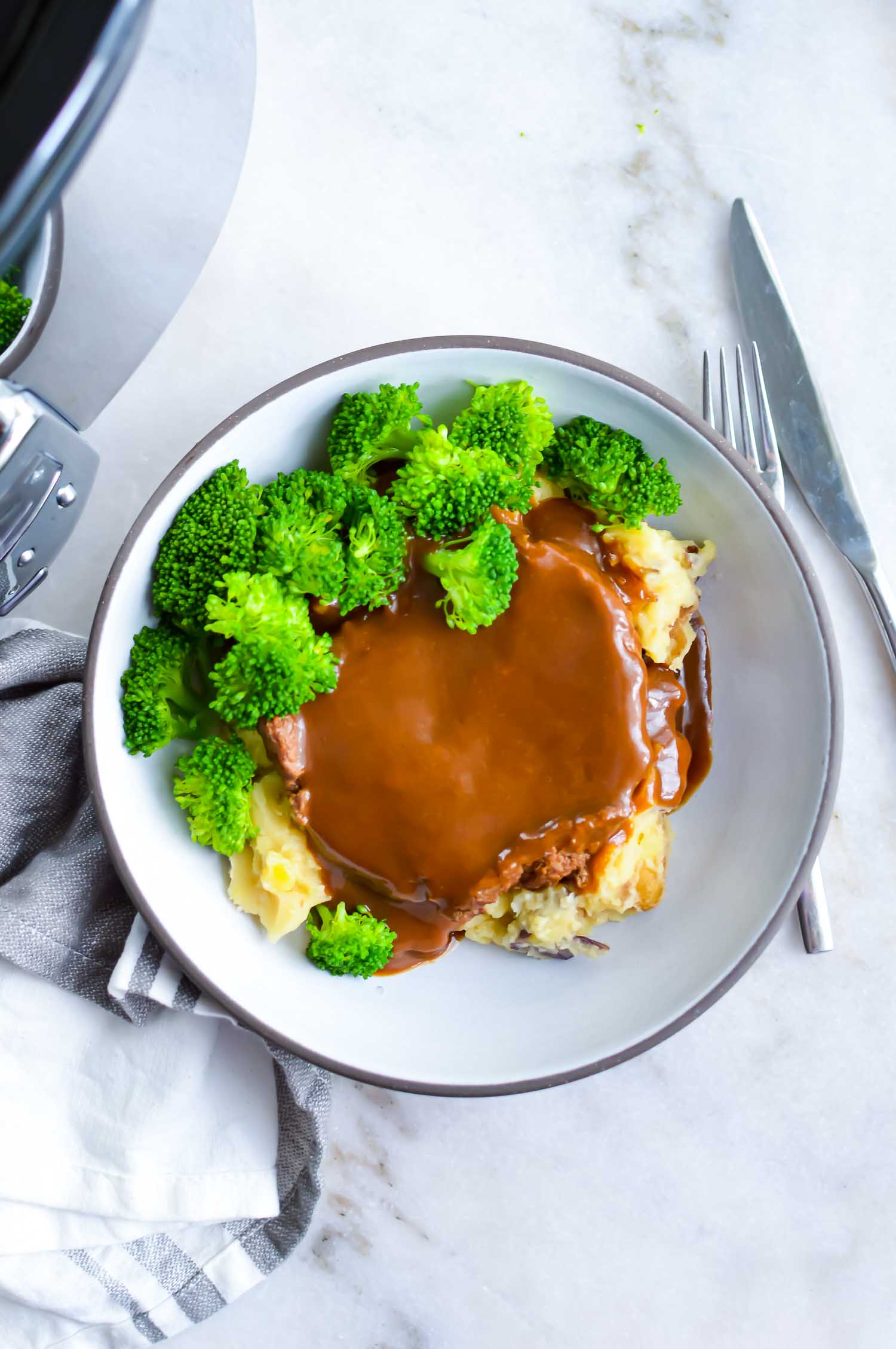 A white bowl with gray rim with cubed steaks, gravy, potato, and broccoli with a fork in it with a fork on the side of it.