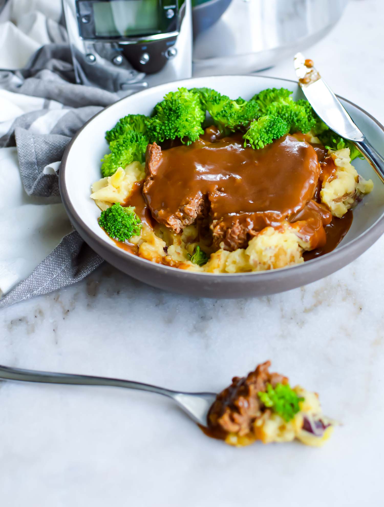 A white bowl with gray rim with meat, gravy, potato, and broccoli with a knife on top with a fork with a bite of food laying in front with a silver slow cooker behind it.