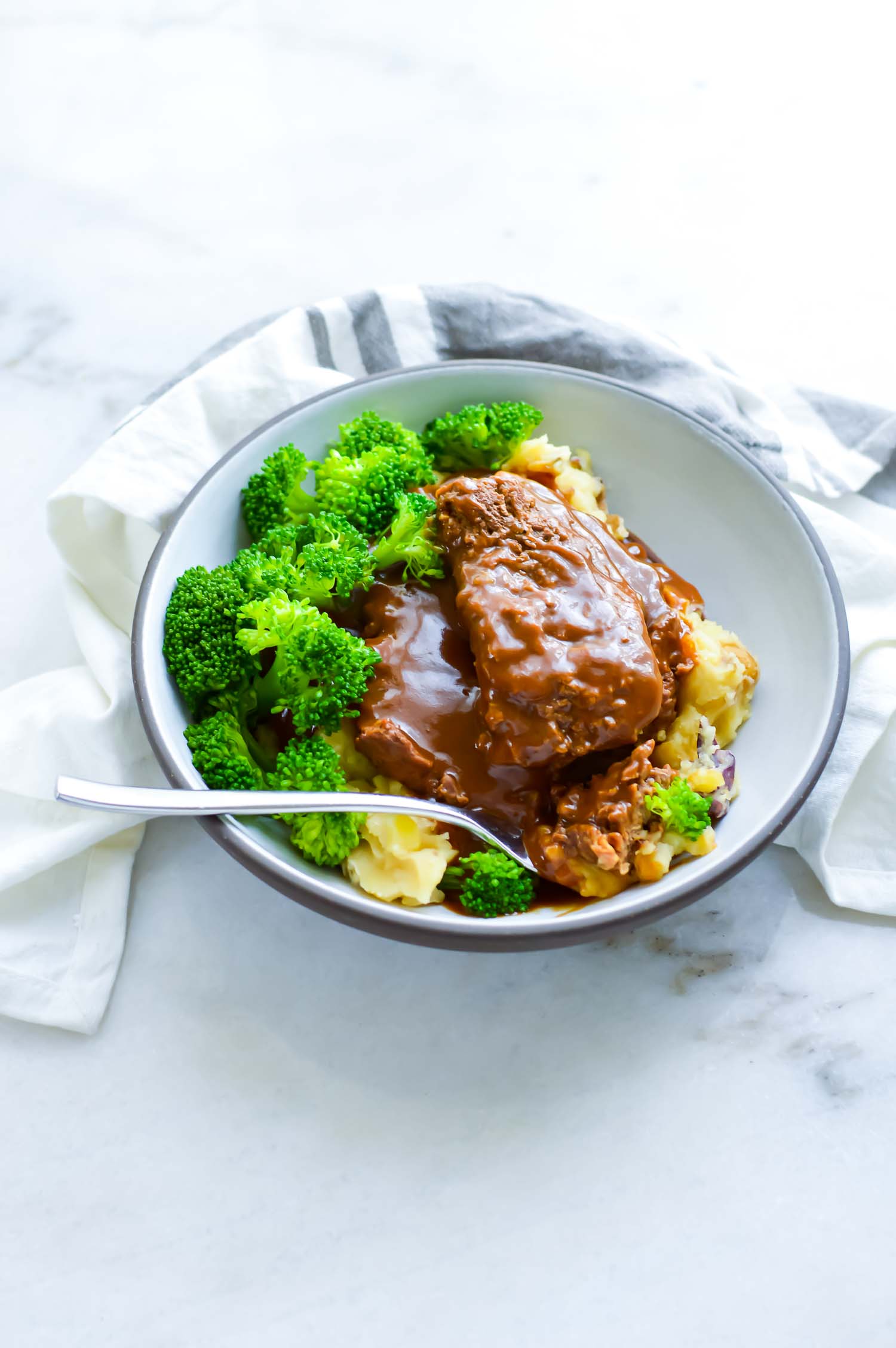A white bowl with gray rim with cubed steaks with gravy, potato, and broccoli with a fork in it.
