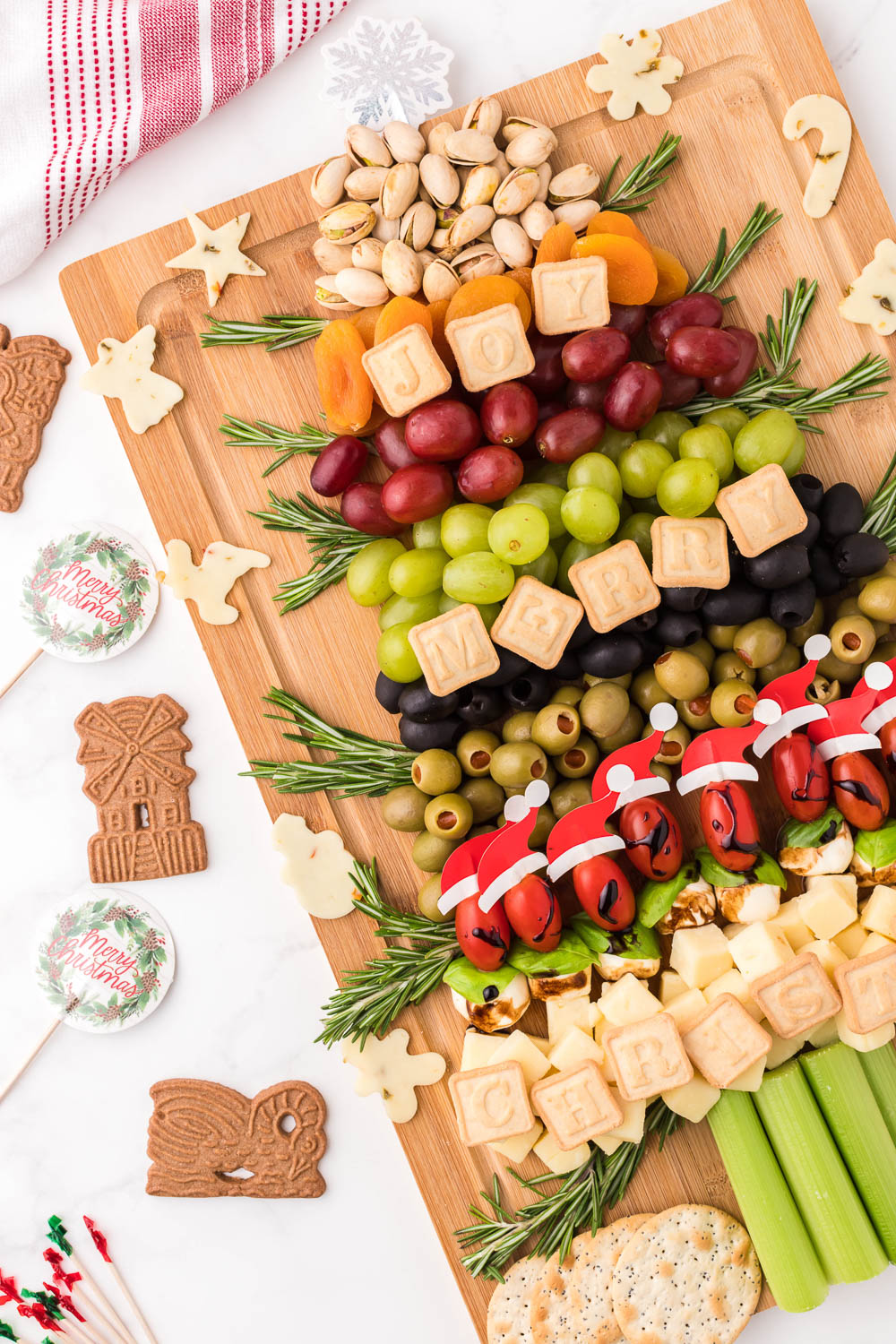 A wooden board at an angle filled with snacks, fruits and food in the shape of a Christmas tree with cookies and signs on the side.
