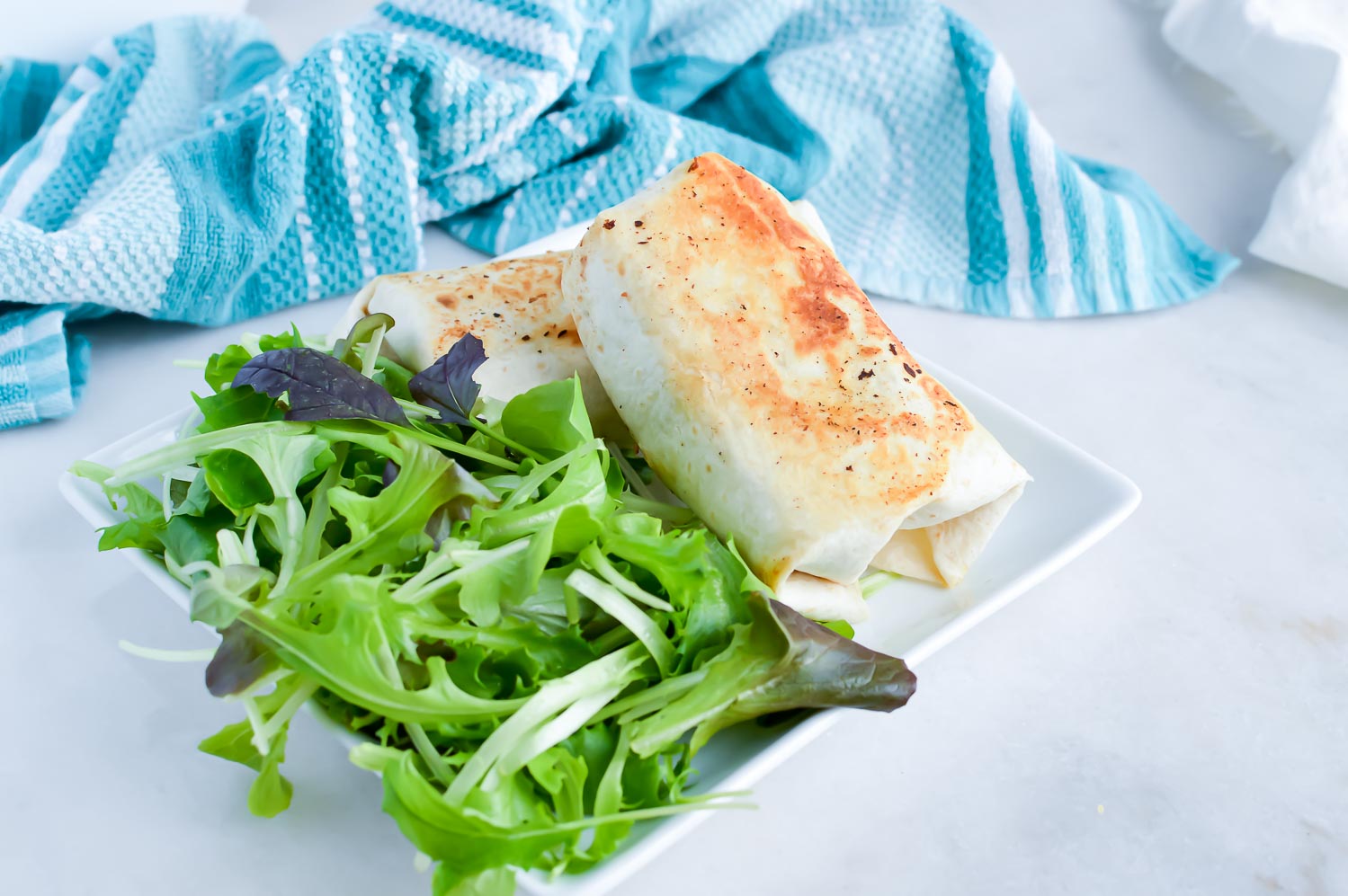 A square white plate with two burritos on top of each other and a green salad with a blue towel at the top.
