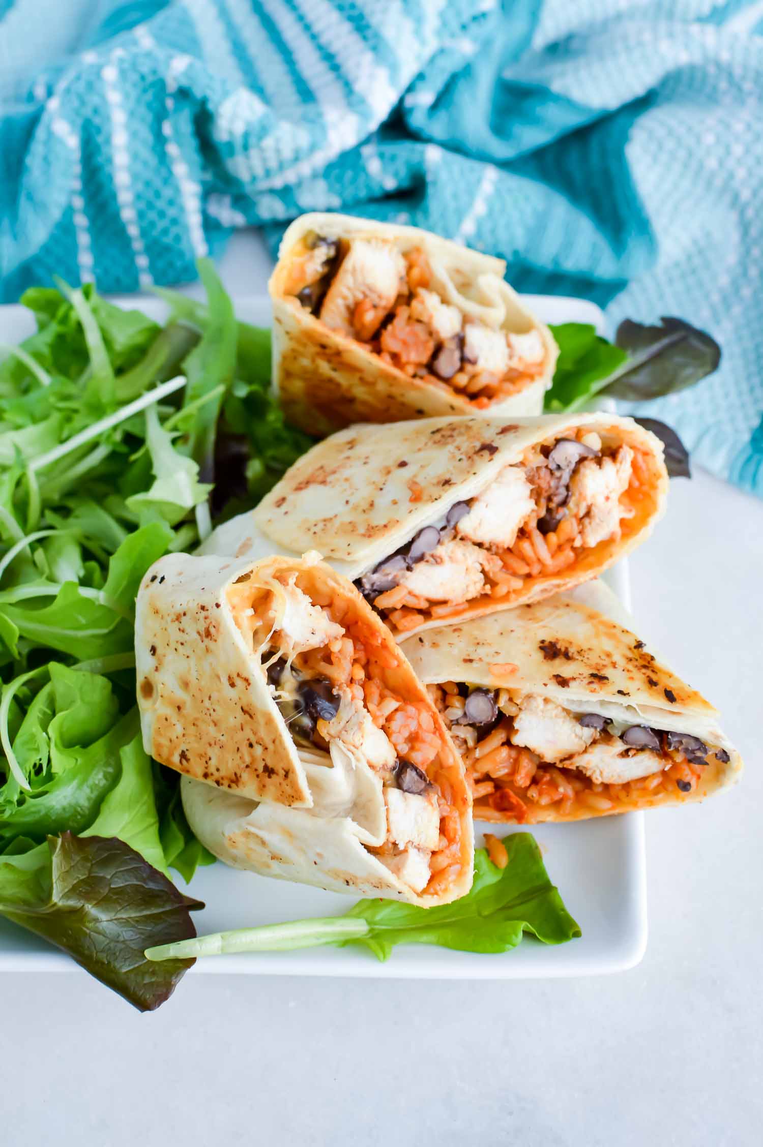 Four cut burritos on a square white plate with a green salad with a blue towel at the top.