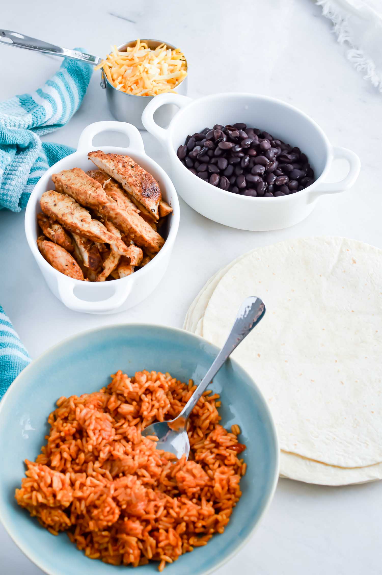White bowls with cooked, sliced chicken, black beans, Spanish rice and a metal measuring cup with shredded cheese in it for the chicken burrito recipe