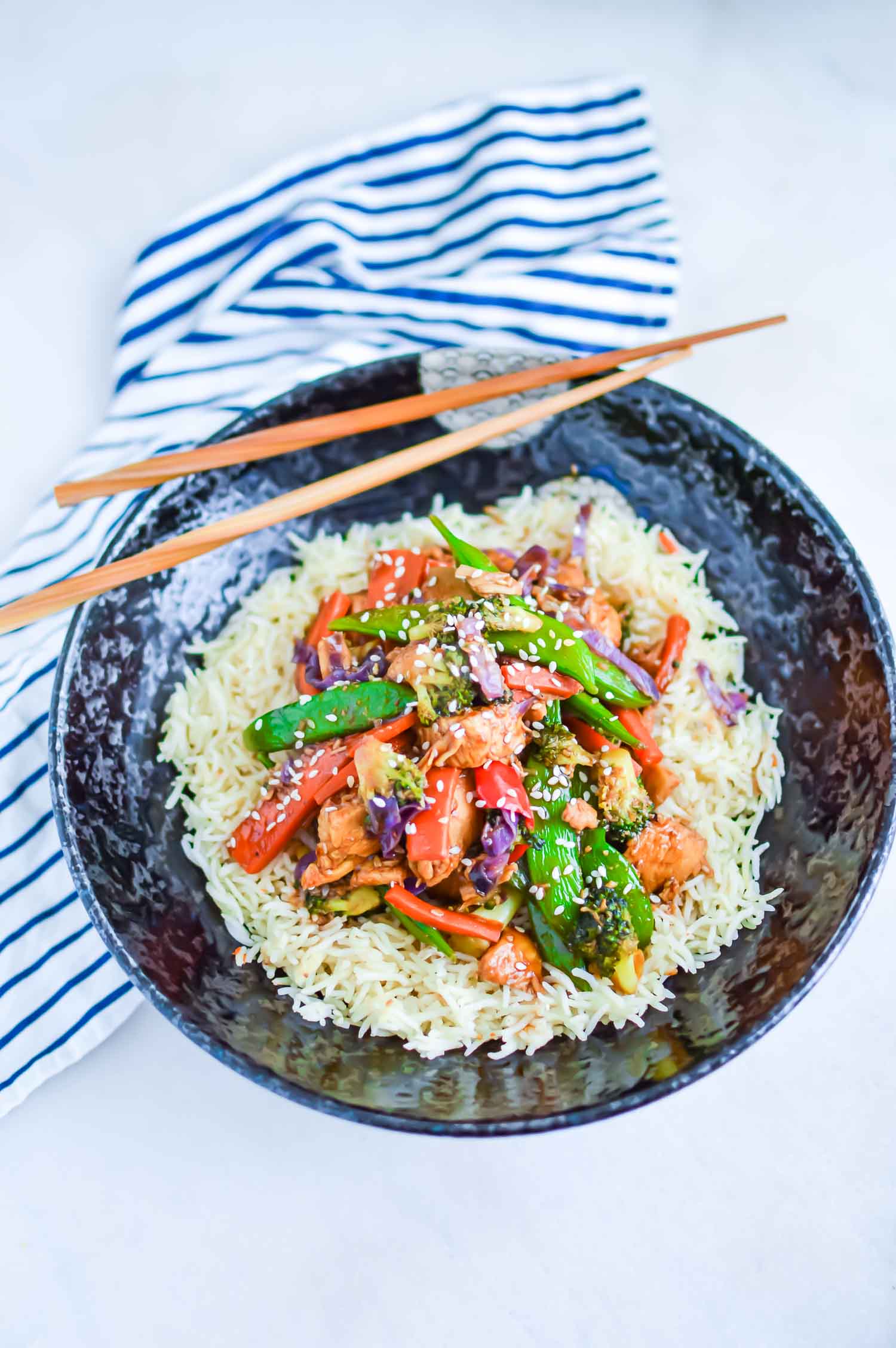 Chicken Stir-fry with vegetables over rice in a bowl with chopsticks  and blue and white striped napkin