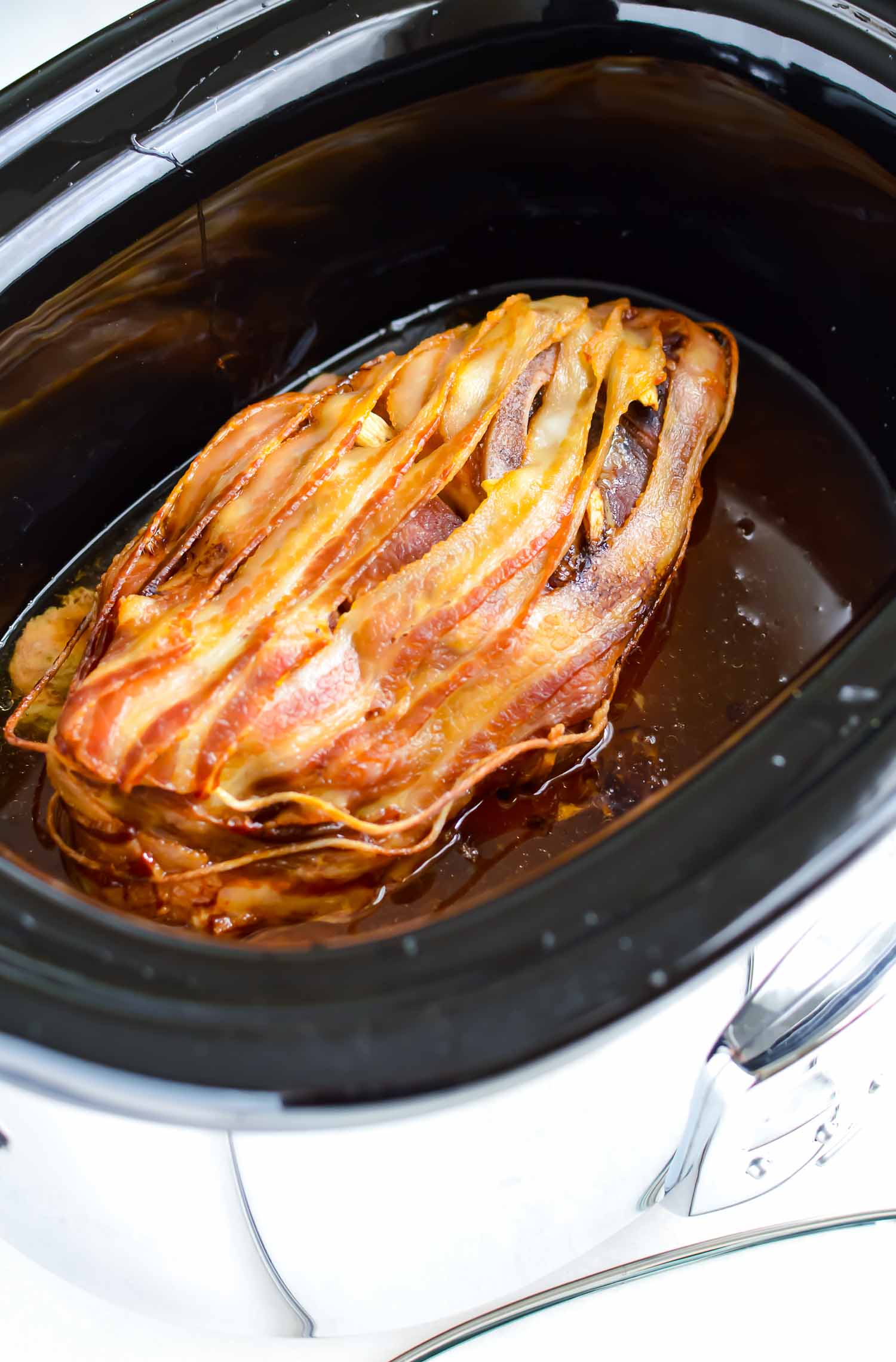 A black crockpot with a pork butt wrapped in bacon.