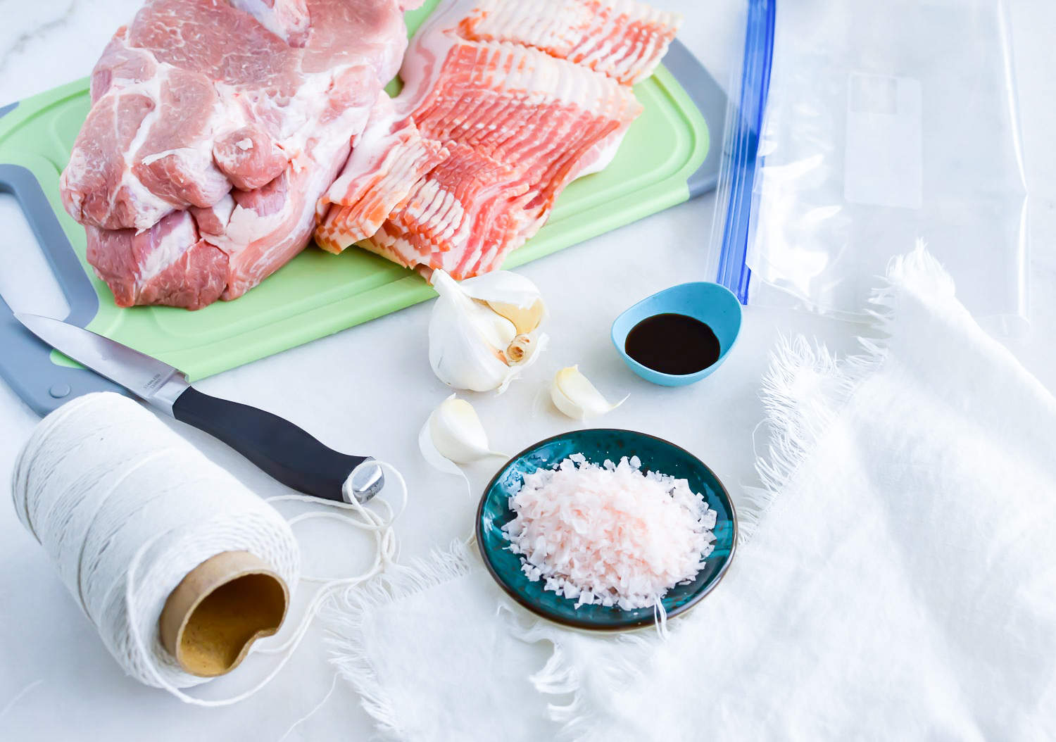 Ingredients for Kalua pork recipe - pork butt, bacon, garlic, salt, twine, and a knife.
