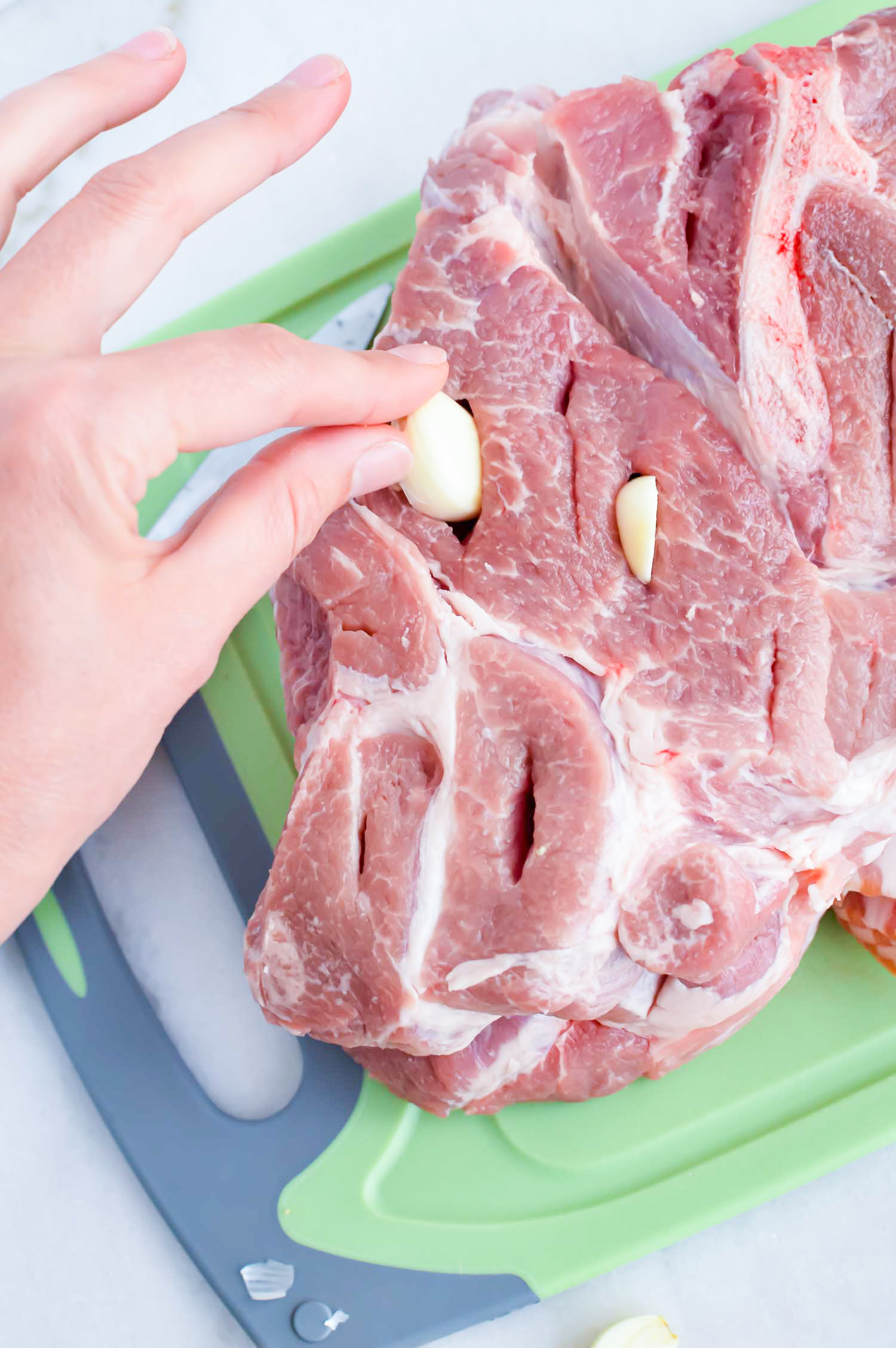 A green cutting board with a pork butt with slits in it with a hand placing garlic pieces in the slits. 