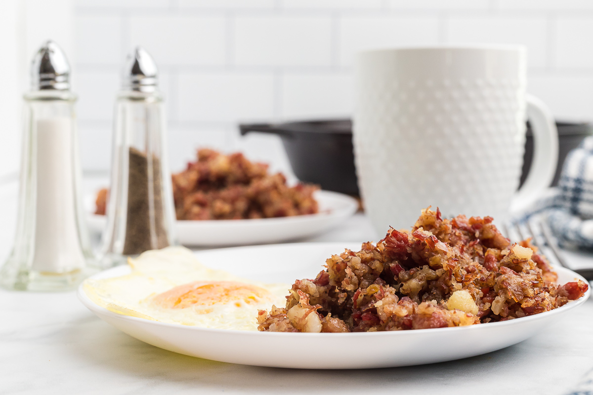 A white plate with hash and an egg on it with salt and pepper shakers and a white mug with a white plate of hash behind it.