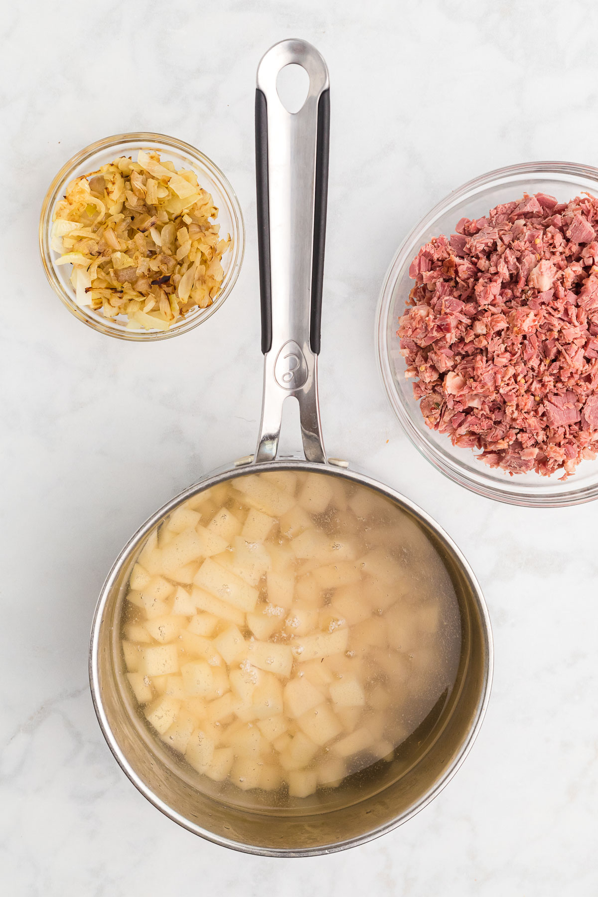 A silver saucepan of water and cut potatoes, two glass bowls: corned beef and cooked onions.