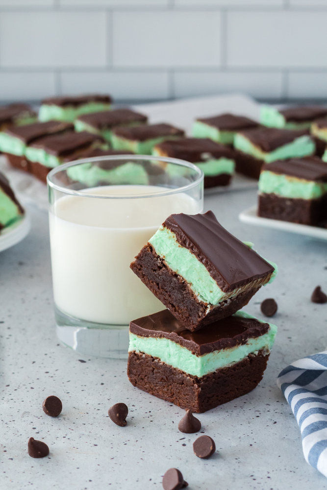 Two chocolate mint brownies stacked on top of each other next to glass of milk with chocolate chips scattered.