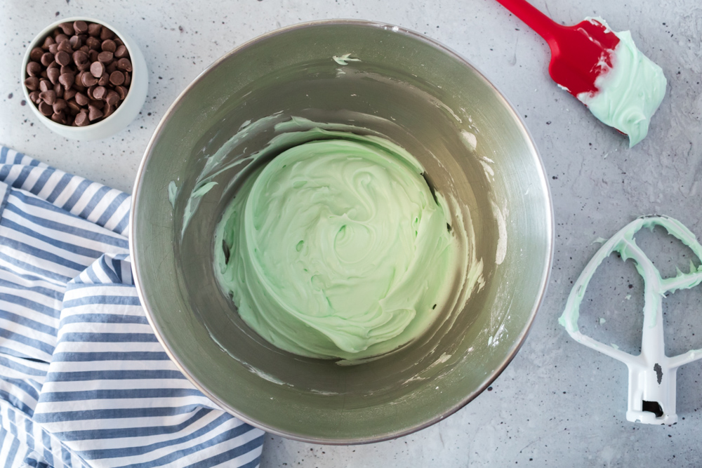 A silver bowl with green frosting in it with a red spatula and a white whisk on the side.
