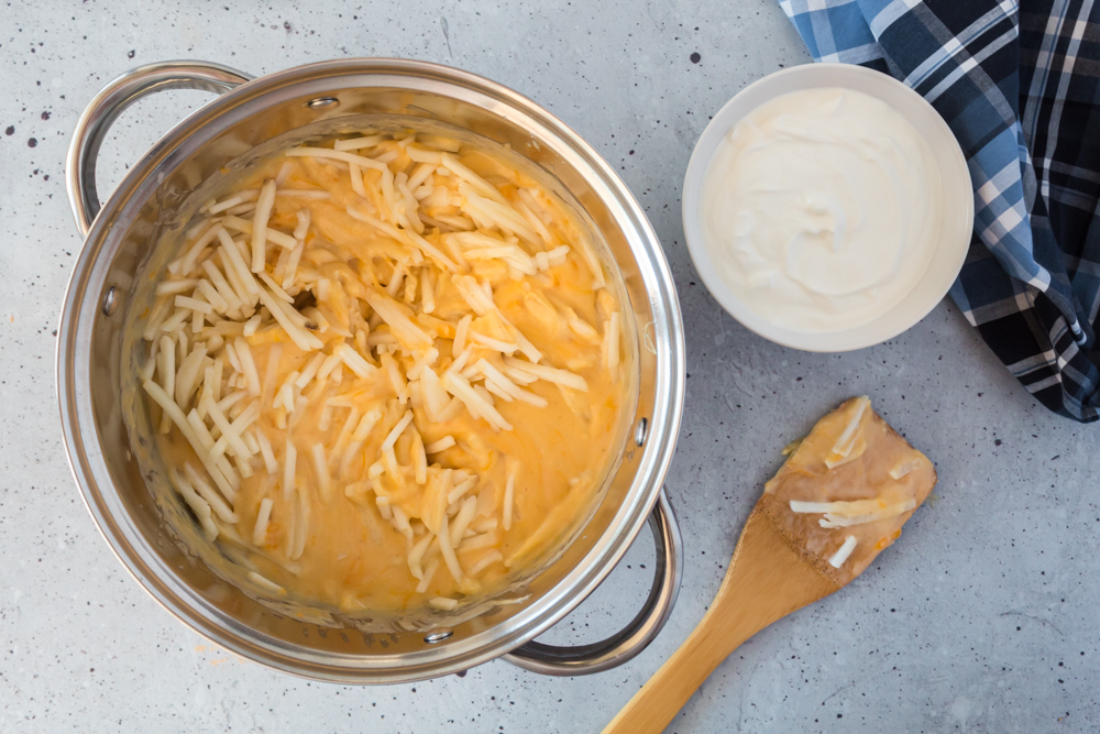 A metal pot with frozen hash browns and a cheese sauce with a white bowl of white liquid on the side with a wooden spoon on the side.