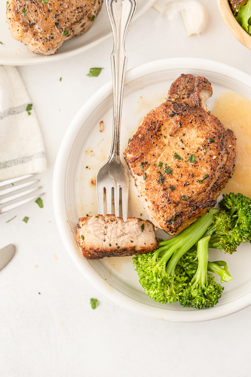 A white plate with a pork chop with a bite out of it with a silver fork, broccoli and applesauce on it with other portions around it.