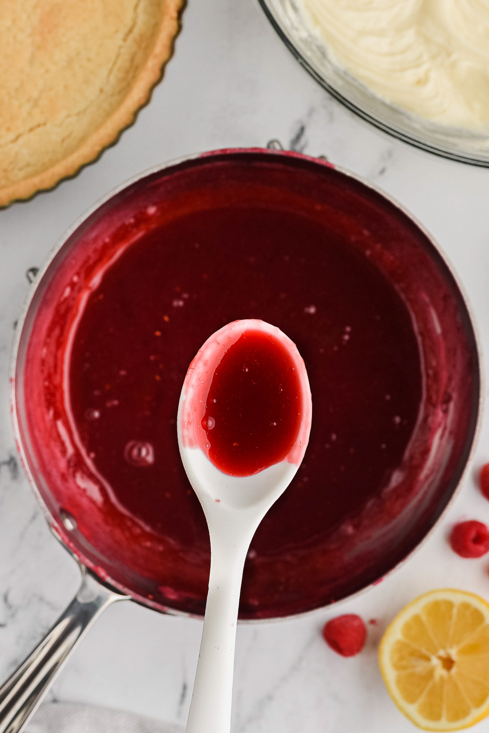 Raspberry filling in a metal pan with a white spoon in it with white filling and a pastry shell on the side.