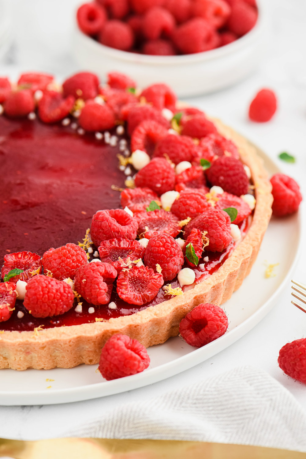 Raspberry fruit tart on a white plate with raspberries, mint leaves and a gold fork.
