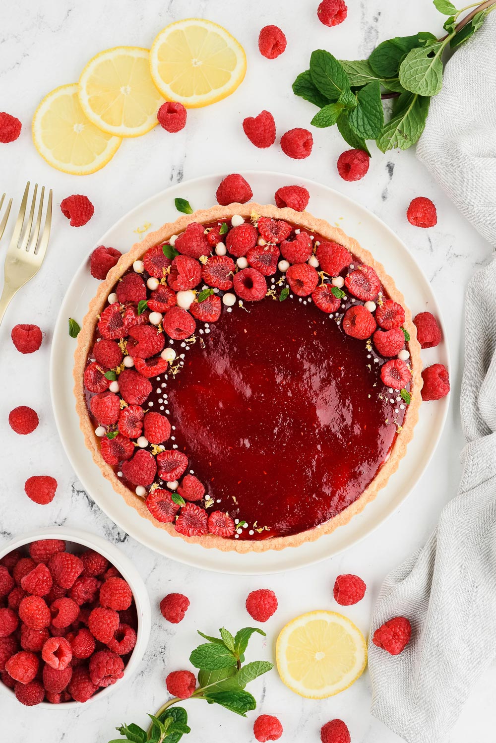 Raspberry fruit tart on a white plate with raspberries, mint leaves, lemon slices and gold forks on the side.
