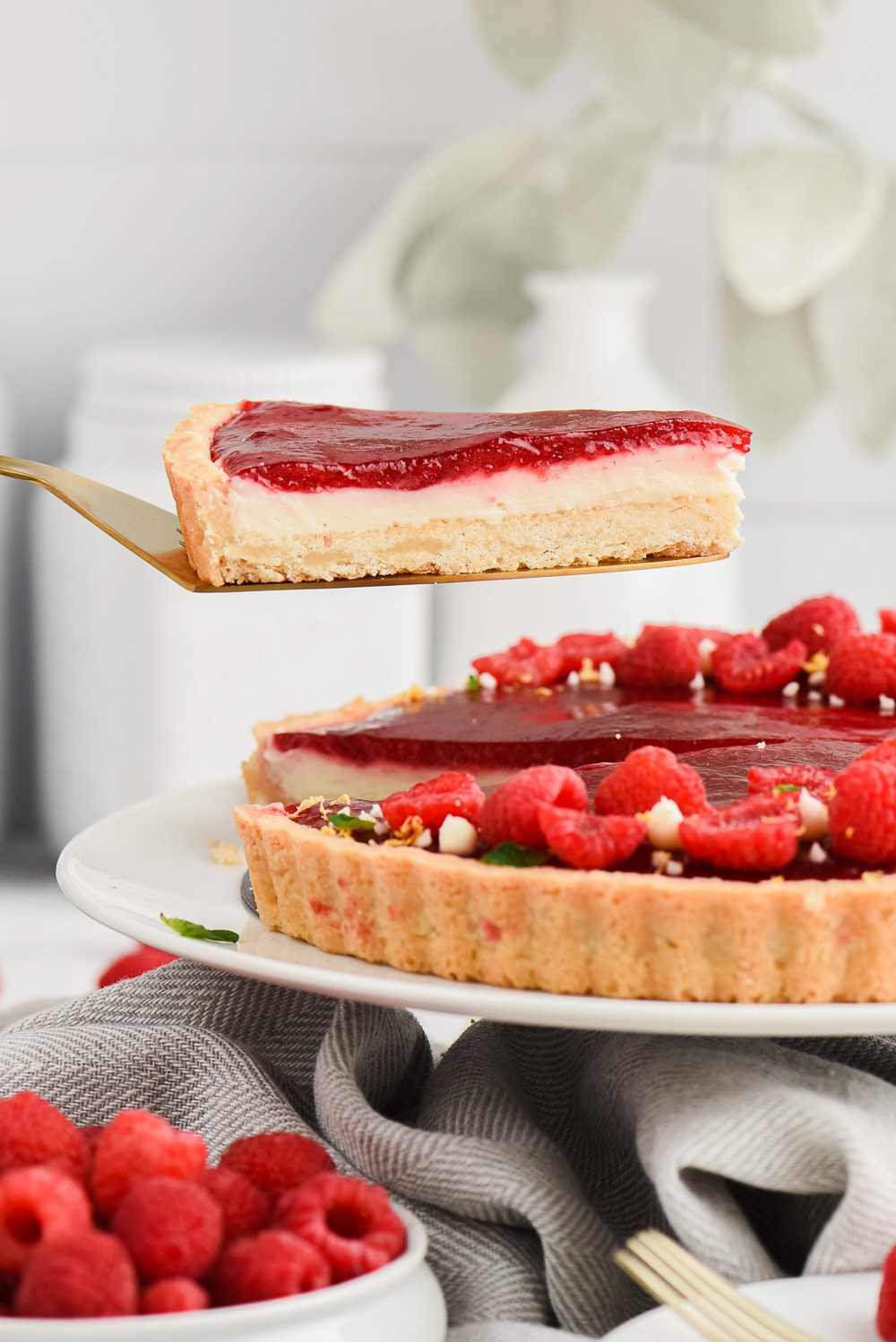 Raspberry fruit tart on a white plate with a slice being raised above it with a golden serving utensil with raspberries on the side.