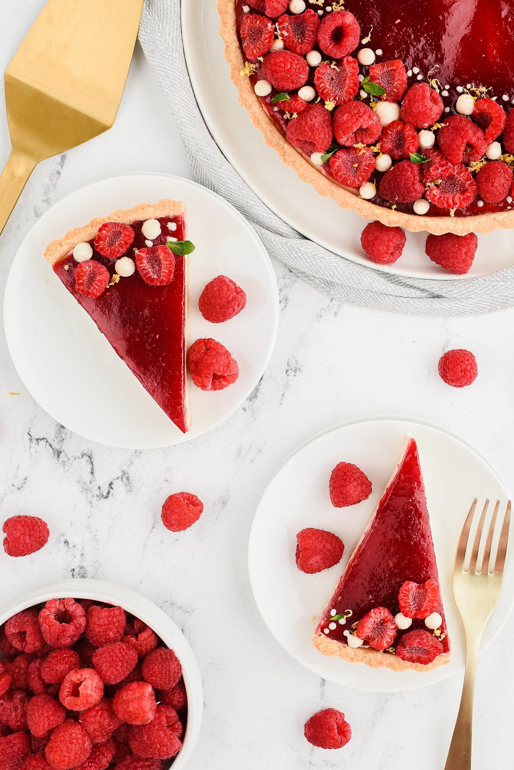 Fruit dessert on a white plate with raspberries on the side. There are two white plates with slices of the tart on them with gold utensils.