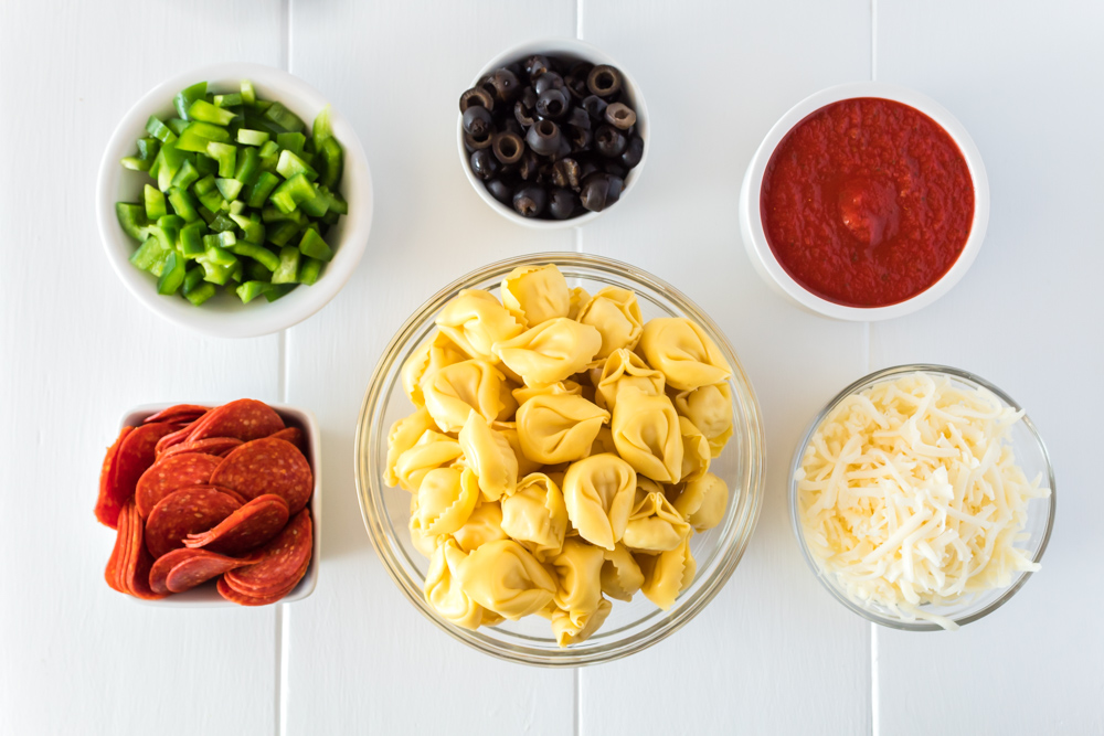 Six individual bowls of pepperoni, tortellini, mozzarella cheese, tomato sauce, olives and green peppers.