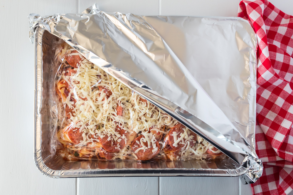 A foil dish with tortellini with a plastic wrap and foil on an angle with a red and white checked towel.