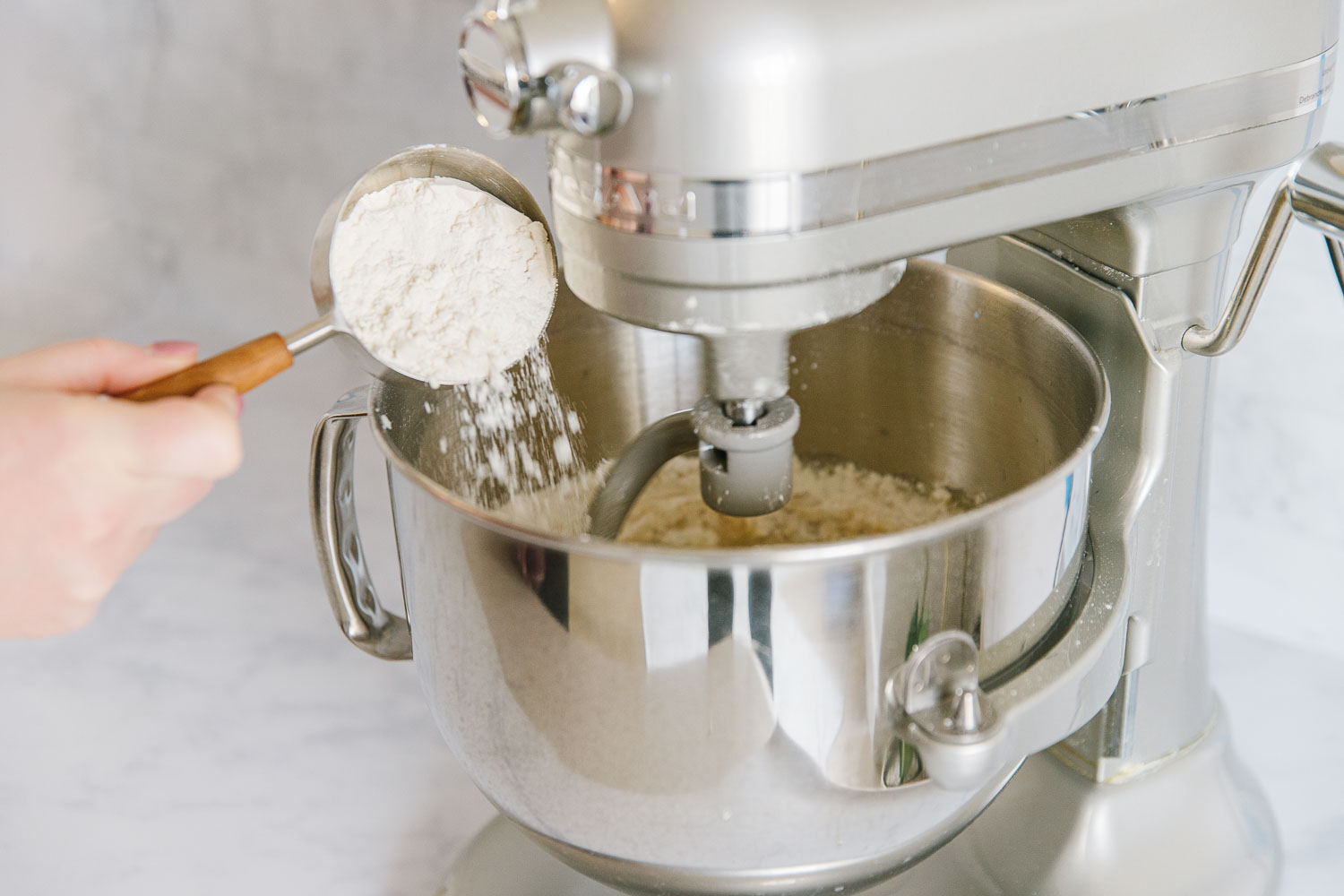 A silver Kitchenaid mixer with a hand adding in a measuring cup of flour into the bowl of ingredients.