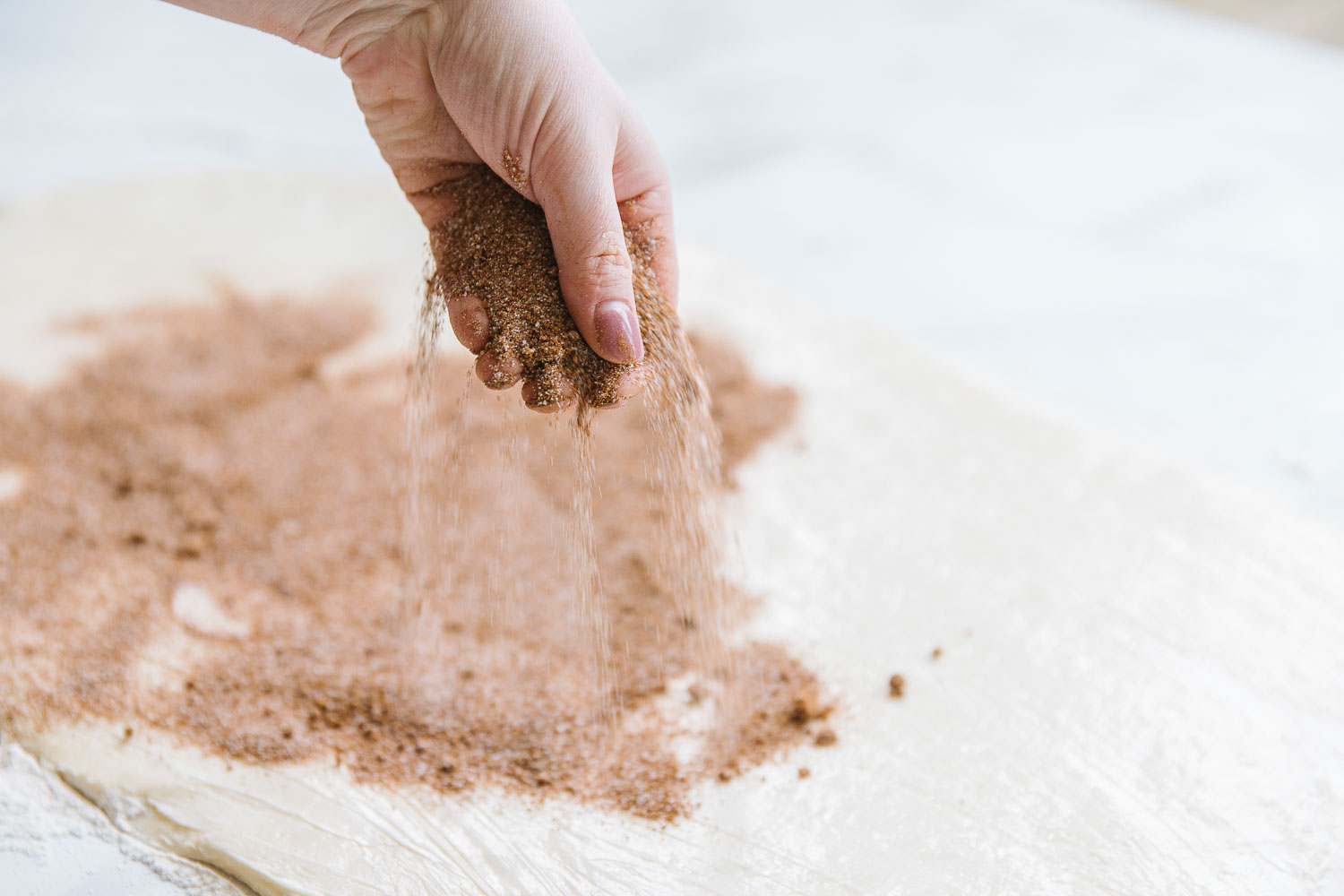 A hand sprinkling cinnamon and sugar mixture onto rolled-out dough.