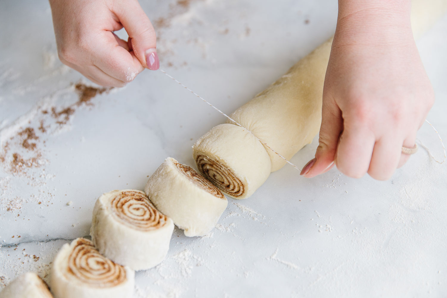 Two hands using floss to cut cinnamon rolls. 