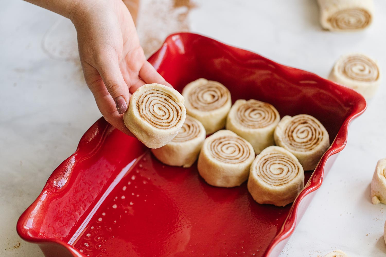 A hand placing raw cinnamon rolls into a red pan where there are 6 cinnamon rolls in the pan.