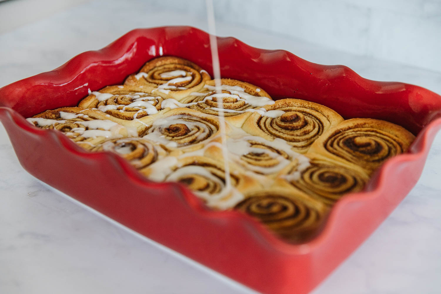 A red pan of cooked cinnamon rolls with icing dripping down onto the rolls.