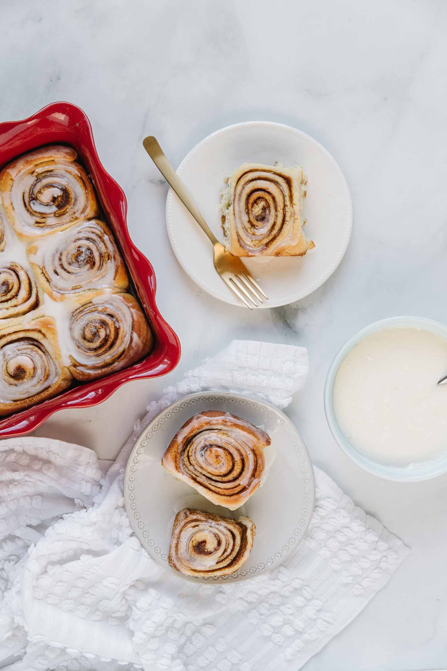 A white plate with two cinnamon rolls with a white bowl of icing, white plate with a cinnamon roll with a gold fork on it and a red pan of other cinnamon rolls.