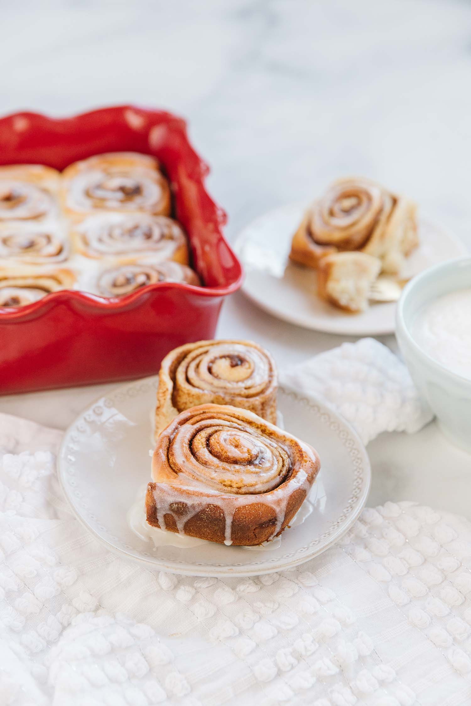 A white plate with two cinnamon rolls with a white bowl of vanilla icing, a white plate with a cinnamon roll and a red pan of other cinnamon rolls.