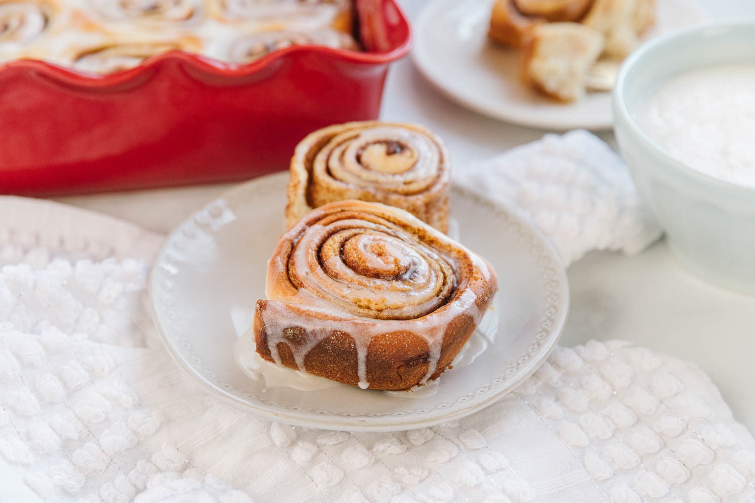 A white plate with two cinnamon rolls with a white bowl of icing, white plate with a cinnamon roll and a red pan of other cinnamon rolls.