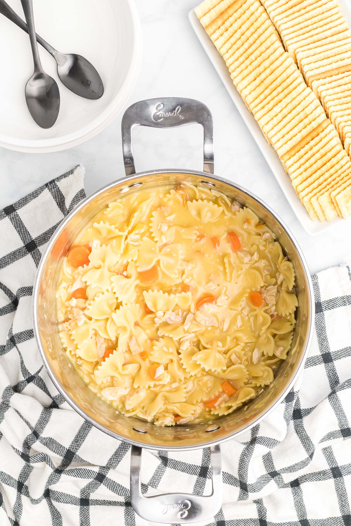 A silver pot with cooked chicken noodle soup with crackers on the side and empty white bowls with two spoons inside. 