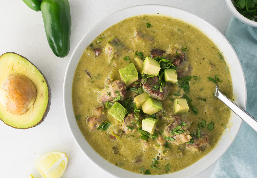 A white bowl of chili verde with cut avocados on top and on the side with a metal spoon.