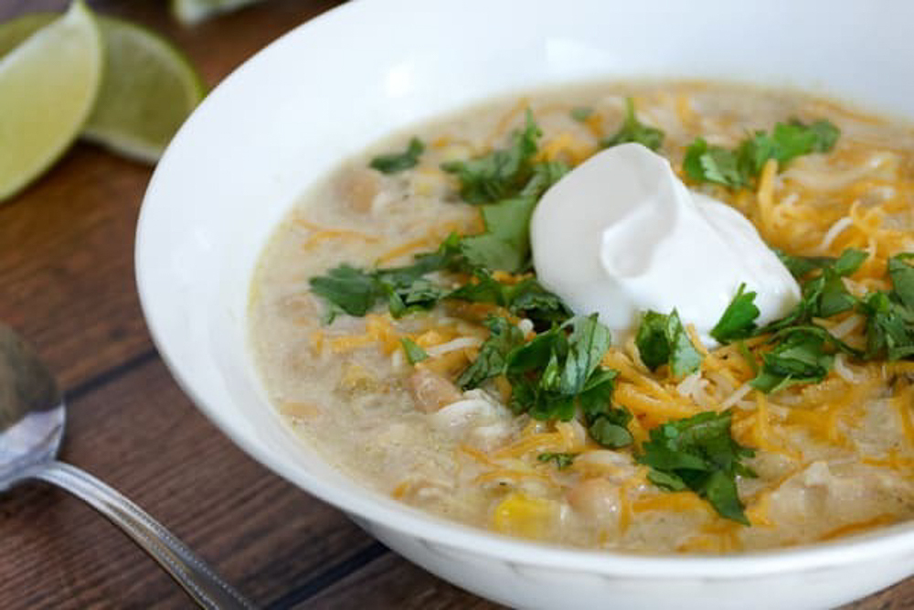 A white bowl of chicken chili with cheese, herbs. and sour cream. 
