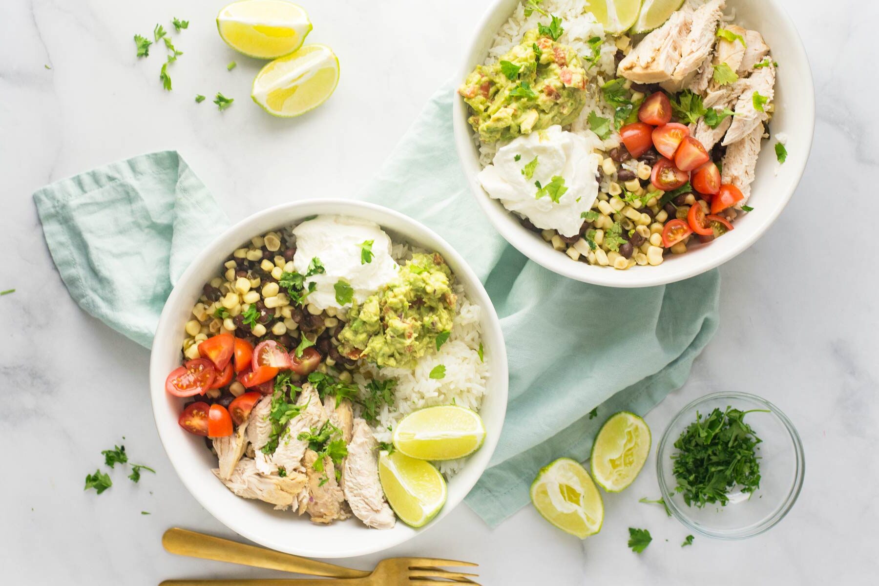 Two white bowls with cilantro lime chicken on top with corn, tomatoes, guacamole and sliced limes and herbs on it.