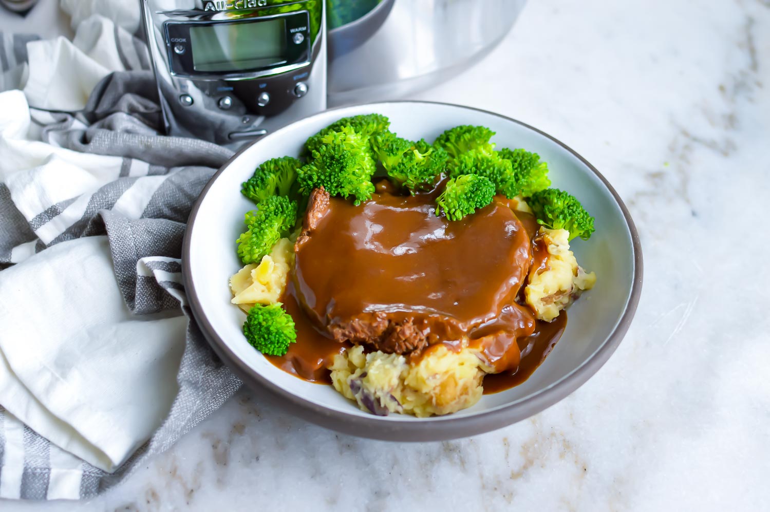 A white bowl with cube steak and gravy and broccoli with a towel on the side.