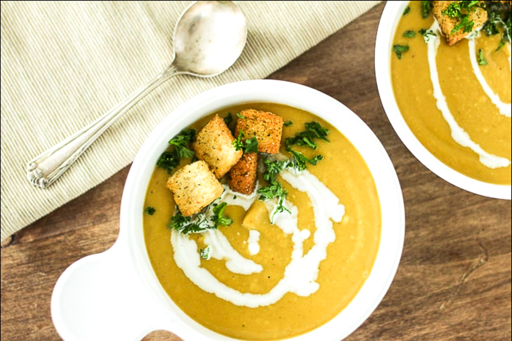 Two white bowls with butternut squash soup in it with a cream drizzle, herbs and croutons inside of it with a silver spoon on the side of it.